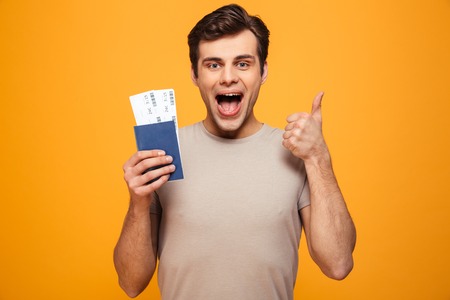 Portrait of a joyful young man holding passport with flying tickets and showing thumbs up isolated...