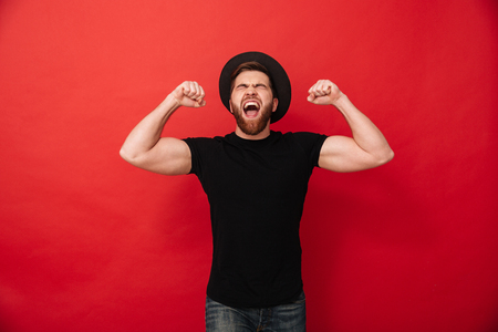 Portrait of ecstatic happy man in black outfit screaming and clenching fists in joy demonstrating his biceps isolated over red backgroundの写真素材