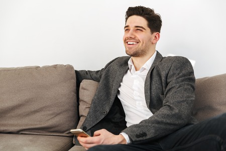 Happy business man sitting on sofa at home while holding smartphone and looking awayの写真素材