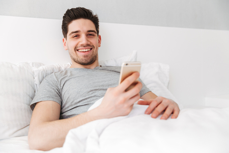Photo of happy awake man in t-shirt smiling while lying alone in bed with white linen and holding golden cell phoneの写真素材