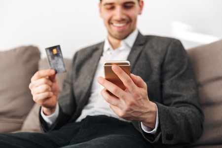 Smiling business man sitting on sofa with credit card at home while using smartphone. Focus on smartphoneの写真素材