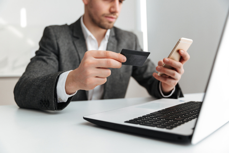 Cropped image of business man sitting by the table with laptop computer while holding credit card and using smartphoneの写真素材