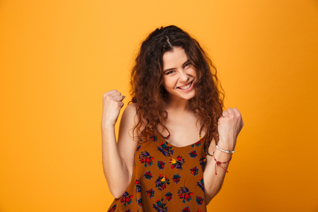 Portrait of a satisfied curly haired girl celebrating success isolated over yellow backgroundの写真素材