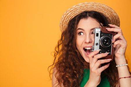 Close up portrait of a young curly haired girl wearing hat and dress making picture with photo camera isolated over yellow backgroundの写真素材