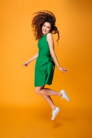 Full length portrait of a cheerful curly haired girl jumping isolated over yellowの写真素材