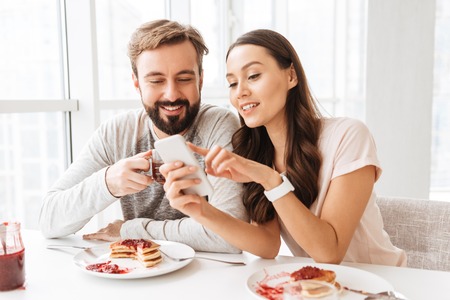 Beautiful young couple having breakfast while sitting together at the table on a kitchen and looking at mobile phoneの写真素材