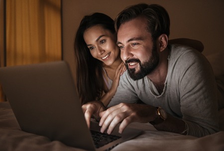 Portrait of a happy young couple using laptop computer while laying on bed at night timeの写真素材