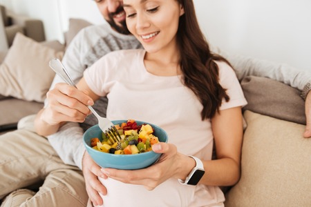 Cropped image of a smiling young pregnant couple relaxing on a couch together, woman having healthy breakfastの写真素材