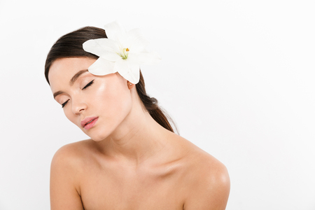Young relaxed asian half-naked lady with closed eyes standing isolated with lily flower on shoulderの写真素材