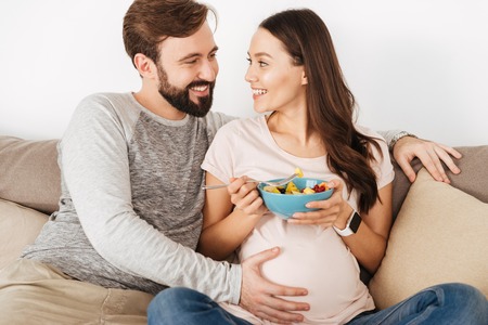 Excited young pregnant couple relaxing on a couch together, woman having healthy breakfastの写真素材