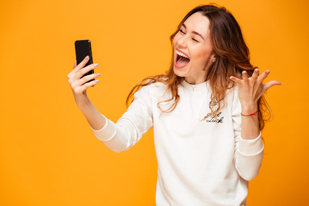 Image of excited emotional young woman standing isolated over yellow background make selfie by mobile phone.の写真素材