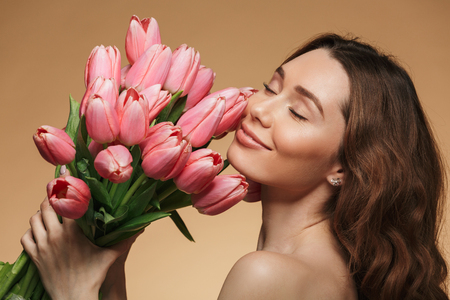 Image in profile of brunette woman with long hair holding and smelling bouquet of pink flowers isolated over beige backgroundの写真素材
