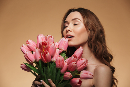 Beautiful woman 20s with natural makeup taking pleasure with closed eyes while holding bouquet of tulips isolated over beige backgroundの写真素材
