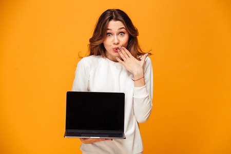 Confused brunette woman in sweater showing blank laptop computer screen while covering her mouth and looking at the camera over yellow backgroundの写真素材