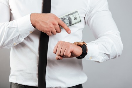 Close up of a businessman holding bunch of money banknotes and pointing finger at his wrist watch isolated over gray backgroundの写真素材