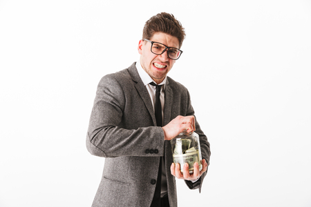 Portrait of a funny young businessman trying to open glass jar full of money banknotes isolated over white backgroundの写真素材