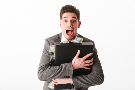 Portrait of a joyful young businessman hugging briefcase full of money banknotes isolated over white backgroundの写真素材
