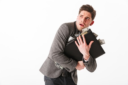 Portrait of a terrified young businessman holding briefcase full of money banknotes isolated over white backgroundの写真素材