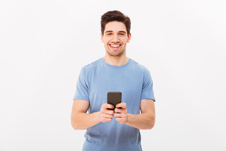 Happy man 30s in casual t-shirt typing sms or browsing internet using mobile phone isolated over white backgroundの写真素材