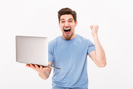 Successful brunette guy in t-shirt clenching fist and rejoicing like lucky guy or winner while using laptop isolated over white backgroundの写真素材