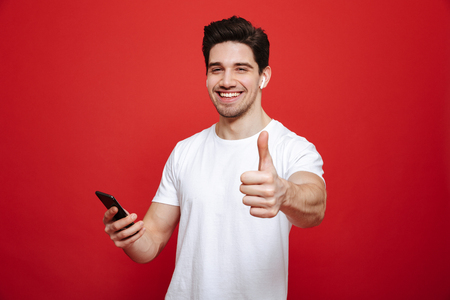 Portrait of an excited young man in white t-shirt listening to music with wireless earphones and mobile phone while showing thumbs up isolated over red backgroundの写真素材