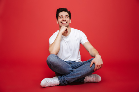 Portrait of a pensive young man in white t-shirt sitting on a floor and looking away at copy space isolated over red backgroundの写真素材