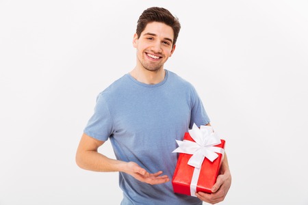 Smiling man in t-shirt presents gift box and looking at the camera over grey backgroundの写真素材