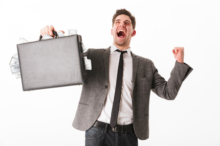 Portrait of a happy young businessman holding briefcase full of money banknotes and celebrating isolated over white backgroundの写真素材