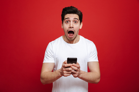 Portrait of a surprised young man in white t-shirt holding mobile phone isolated over red backgroundの写真素材