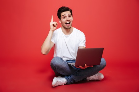 Portrait of a confident young man in white t-shirt holding laptop computer while sitting on a floor and pointing finger up isolated over red backgroundの写真素材