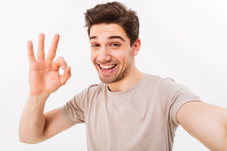 Cheerful man in casual t-shirt and bristle on face smiling on camera with alright sign while taking selfie isolated over white backgroundの写真素材