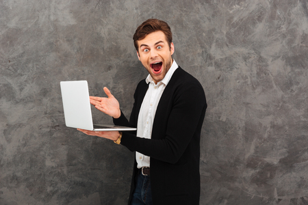 Portrait of a happy young man dressed in shirt and jacket holding laptop computer isolated over gray backgroundの写真素材