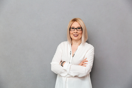 Pleased middle-aged blonde woman in shirt and eyeglasses posing with crossed arms and looking at the camera over grey backgroundの写真素材