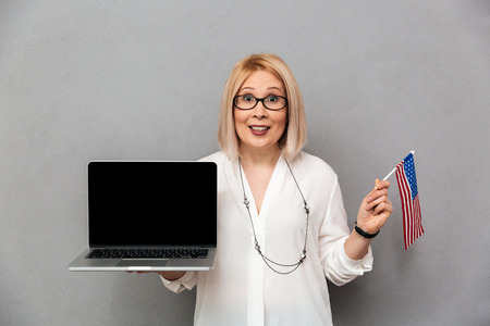 Happy middle-aged blonde woman in shirt and eyeglasses showing blank laptop computer screen while holding USA flag and looking at the camera over grey backgroundの写真素材