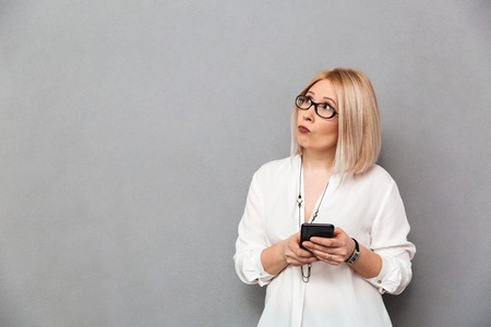 Thoughtful middle-aged blonde woman in shirt and eyeglasses holding smartphone and looking away over...