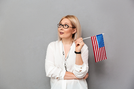 Middle-aged blonde woman in shirt and eyeglasses holding USA flag and looking away over grey backgroundの写真素材