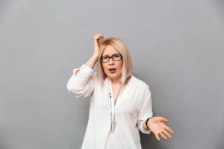 Confused middle-aged blonde woman in shirt and eyeglasses touching her head and looking at the camera over grey backgroundの写真素材
