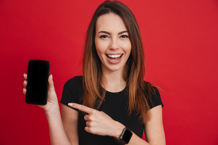 Closeup photo of cheerful adult girl smiling and demonstrating black screen of smartphone with finger isolated over red backgroundの写真素材