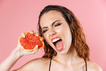 Close up fashion portrait of a screaming young woman wearing makeup dressed in bodysuit squeezing grapefruit while standing isolated over pink backgroundの写真素材