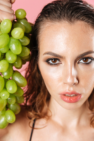 Close up fashion portrait of a seductive young woman wearing makeup dressed in bodysuit posing with grapes while standing isolated over pink backgroundの写真素材
