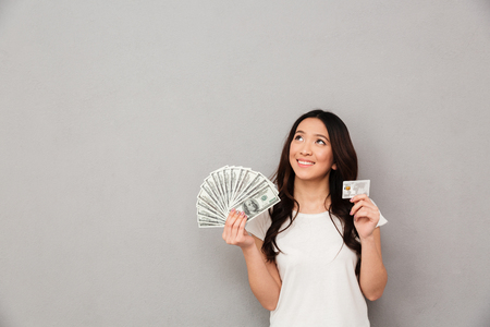 Image of asian content woman 20s holding fan of money dollar banknotes and credit card and looking on copyspace isolated over gray backgroundの写真素材
