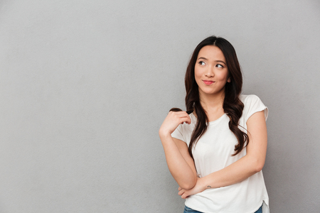 Portrait of asian young woman with playful look posing on camera and looking aside on copyspace isolated over gray background