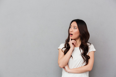 Portrait of chinese surprised woman with dark curly hair posing on camera and looking aside on copyspace with finger on lips isolated over gray backgroundの写真素材