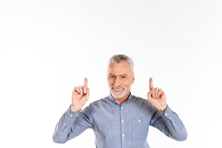 Smiling mature man in blue shirt pointing up with fingers and looking camera isolated over whiteの写真素材