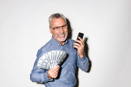 Laughing mature lucky businessman in formal wear and glasses holding smartphone and fan of dollars isolated over whiteの写真素材