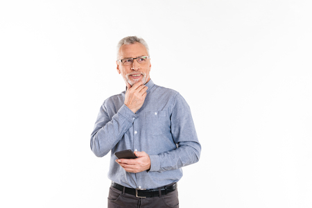 Pensive smiling mature man in glasses holding smartphone and thinking while looking aside isolated over whiteの写真素材