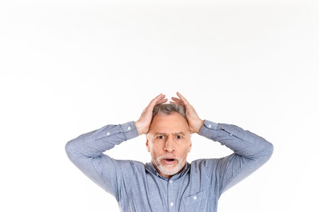 Shocked mature man looking camera with opened mouth and holding his head while posing isolated over whiteの写真素材