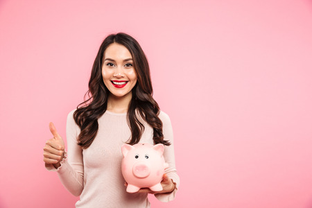 Lovely satisfied woman 20s with long dark hair holding piggybank and gesturing thumb up isolated over pink backgroundの写真素材