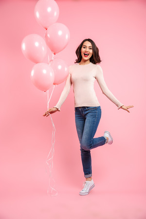 Full length image of wonderful woman 20s in casual clothing looking on camera with delight while holding bunch of air balloons in hand isolated over pink backgroundの写真素材