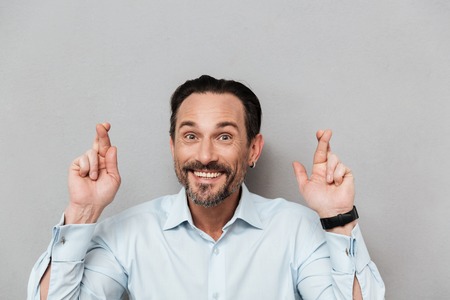 Portrait of a smiling mature man dressed in shirt holding fingers crossed for good luck over gray...